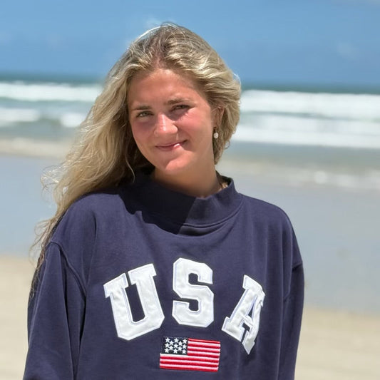 Person wearing a navy blue sweatshirt with 'USA' and an American flag on a beach.
