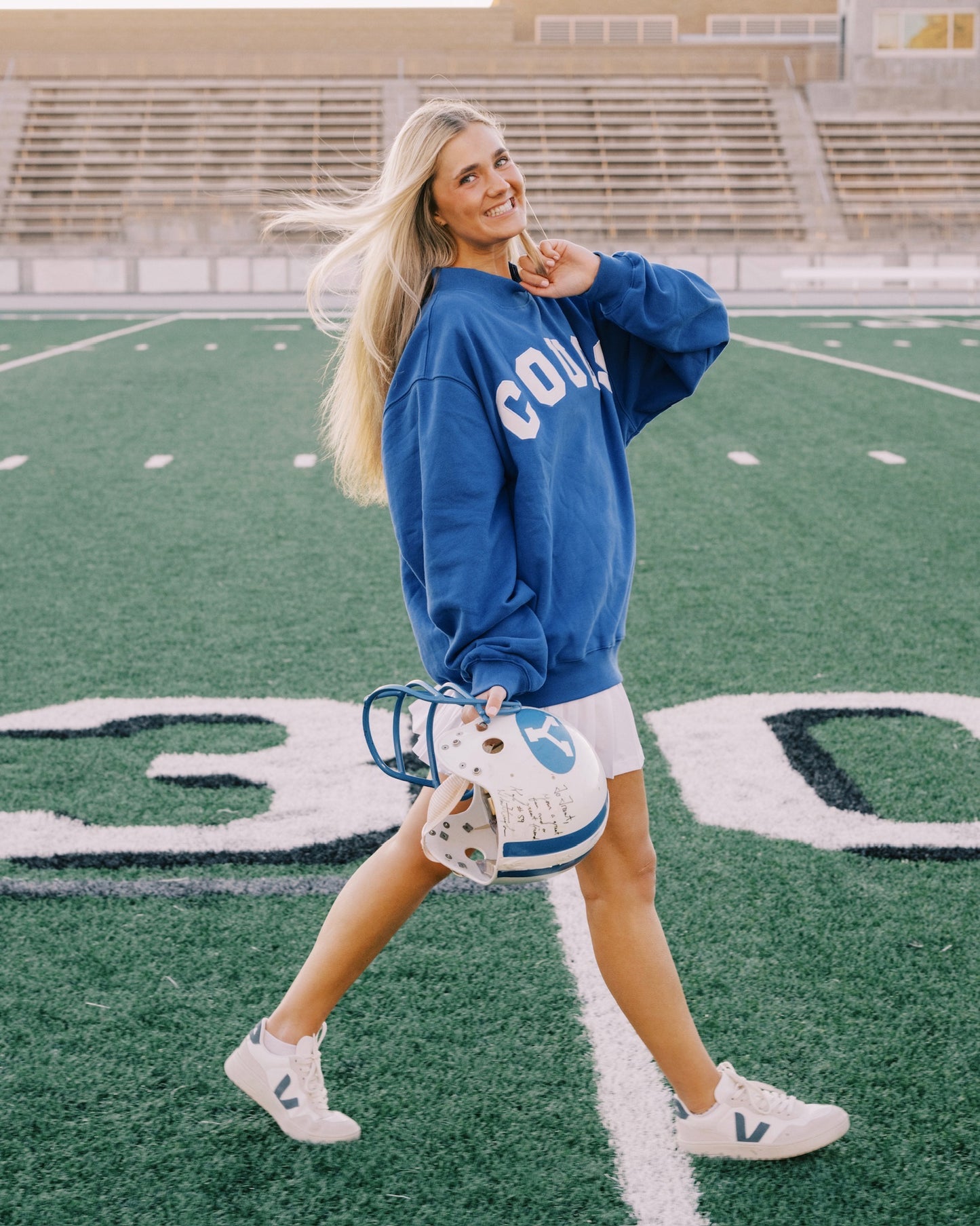 Woman in blue sweatshirt and white shorts standing on a football field holding a football.