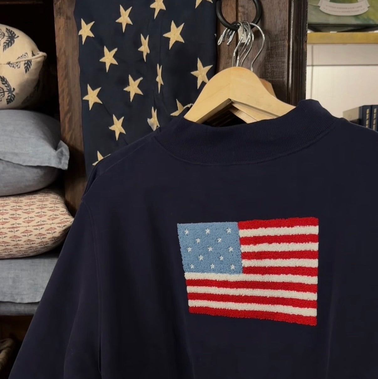 Navy sweatshirt with American flag patch on a wooden hanger against a decorative wall.