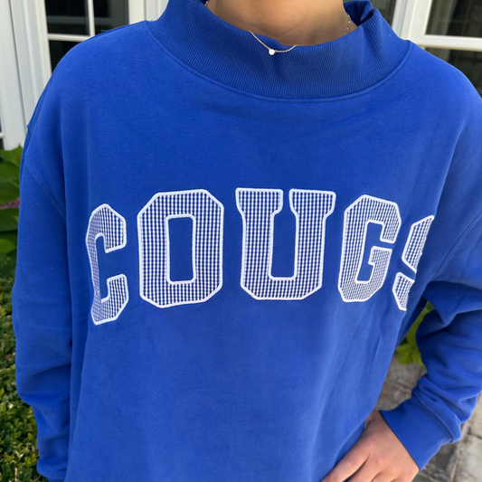 A person wearing a royal blue mock neck sweatshirt with 'COUGS' printed across the chest in white outlined block letters, photographed outdoors near a building entrance.