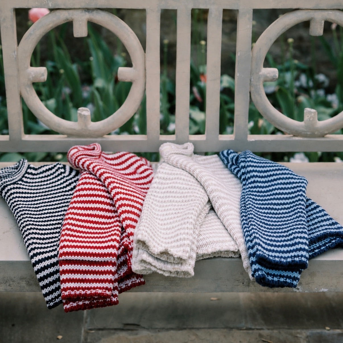 Four knitted sweaters in black and white, red and white, beige, and blue and white stripes on a stone bench with a garden background.