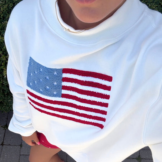 White sweatshirt with American flag design worn by a person on a paved surface.