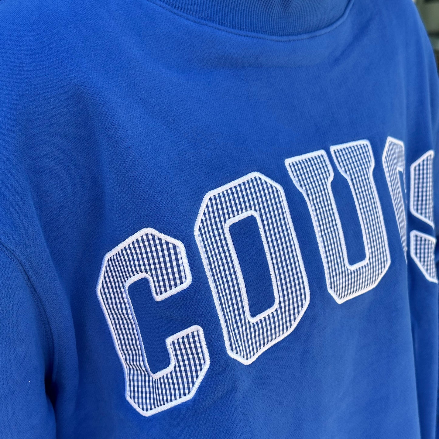 A close-up of a royal blue crewneck sweatshirt featuring the word 'COUS' in large white varsity-style block letters with a grid pattern fill.