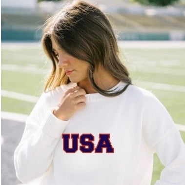Woman wearing a white sweatshirt with 'USA' printed on it, standing outdoors.