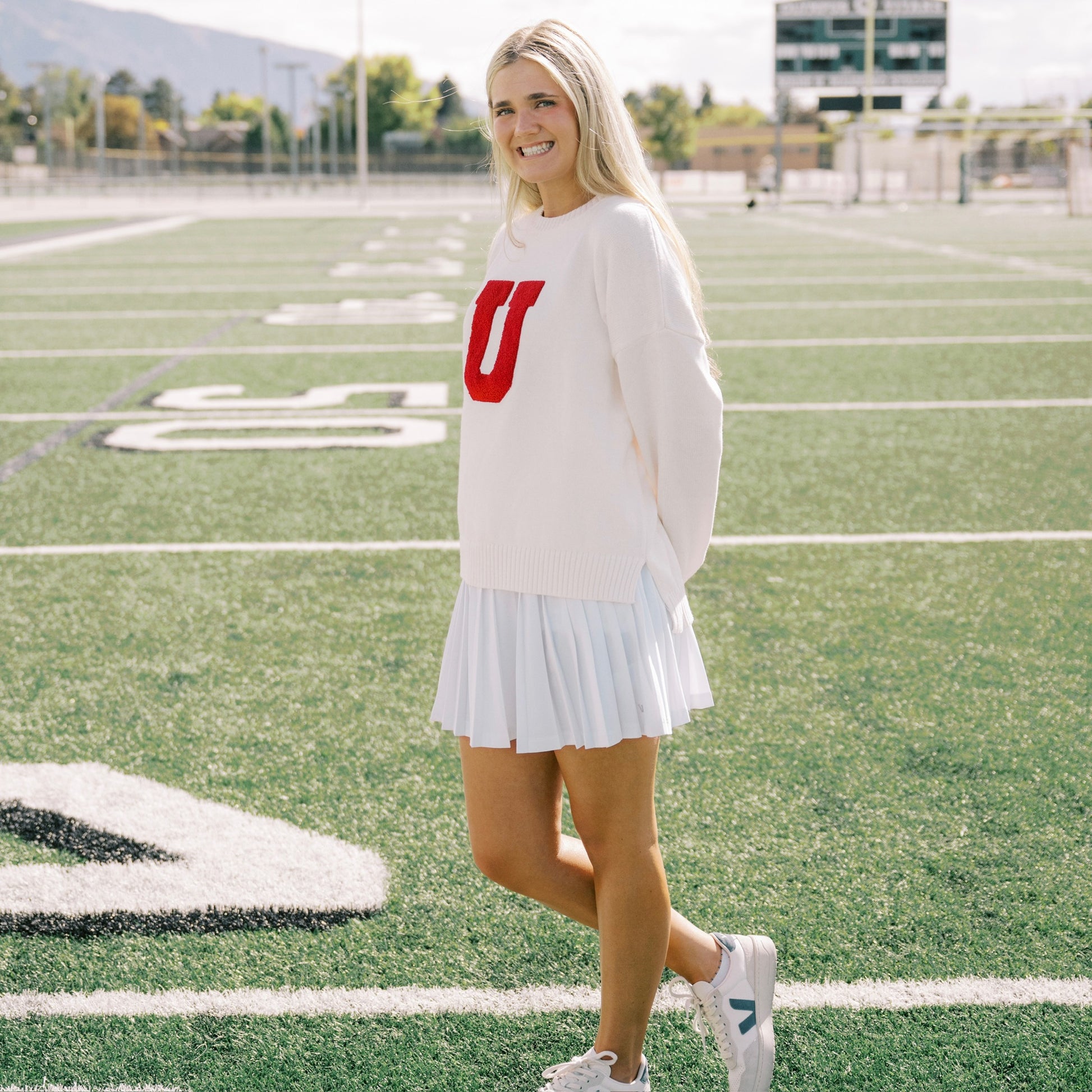 Woman in a white sweater with a red 'U' and white skirt standing on a football field.