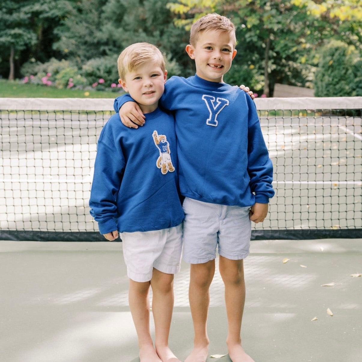 Two young boys in blue sweatshirts standing on a tennis court.